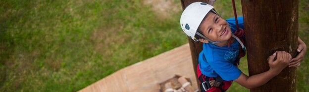 The image shows a young boy wearing a helmet and climbing a tree. He is smiling and appears to be enjoying himself. The background is a grassy area.